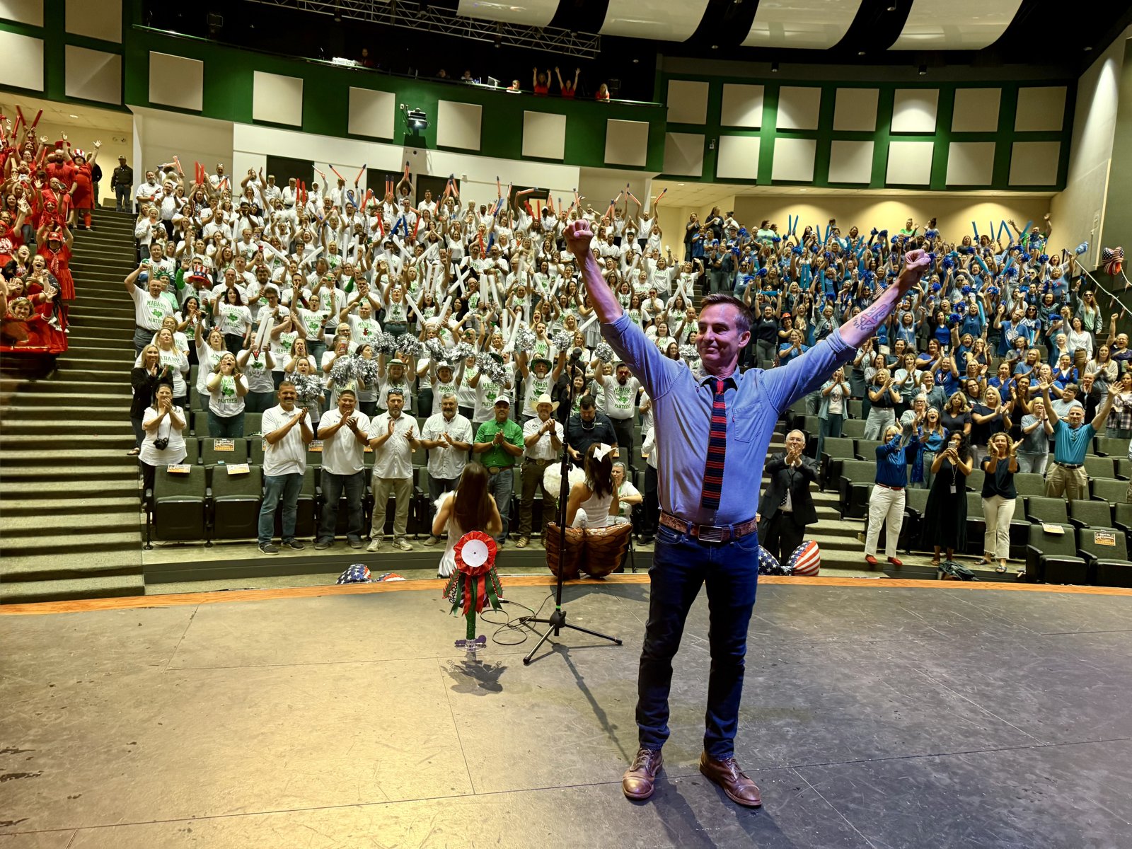 Keni Thomas on stage, arms raised, in front of a cheering school auditorium